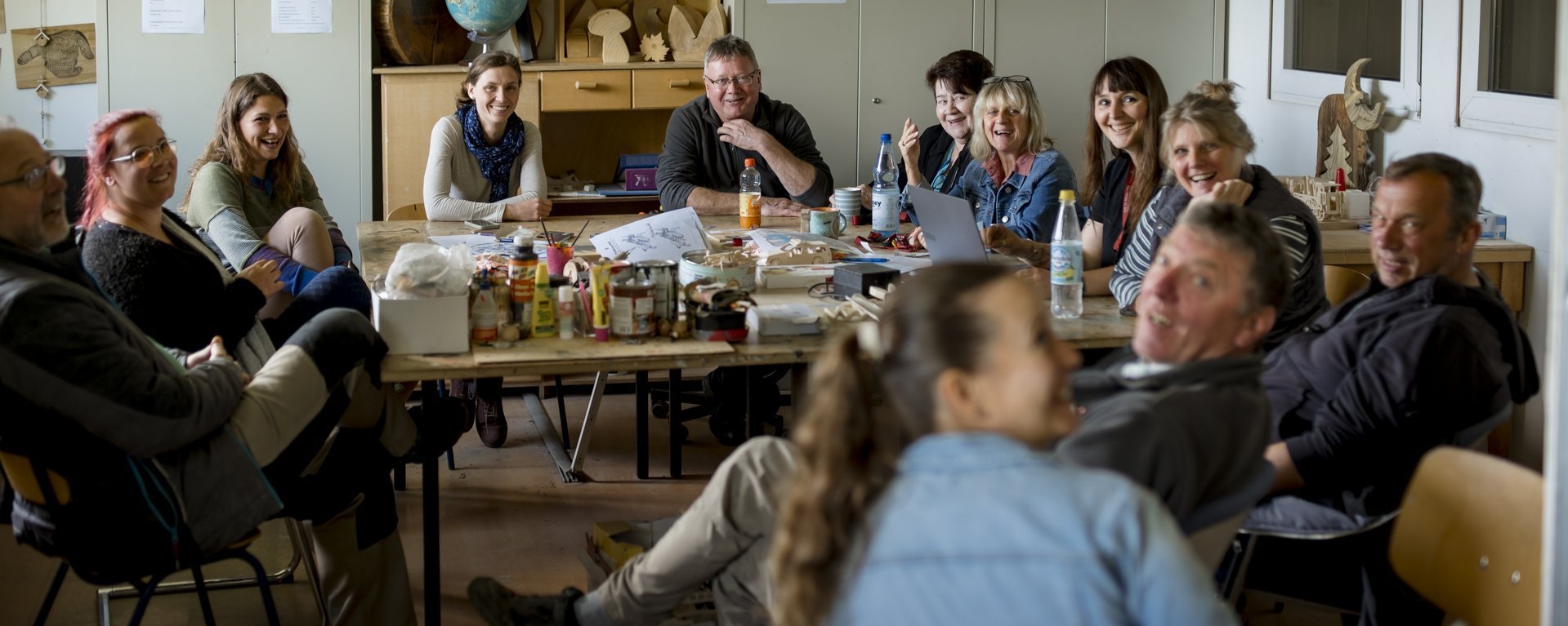 12 Menschen sitzen in einer Werkstatt um einen Tisch, unterhalten sich und lachen..