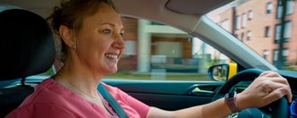 Eine lächelnde Frau in rosa medizinischer Kleidung fährt ein Auto. Sie hält das Lenkrad, trägt einen Sicherheitsgurt und blickt auf die Straße. Im Hintergrund sind unscharfe Wohngebäude zu erkennen.