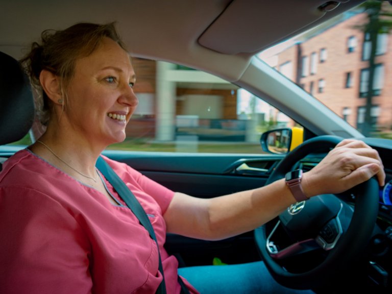 Eine lächelnde Frau in rosa medizinischer Kleidung fährt ein Auto. Sie hält das Lenkrad, trägt einen Sicherheitsgurt und blickt auf die Straße. Im Hintergrund sind unscharfe Wohngebäude zu erkennen.