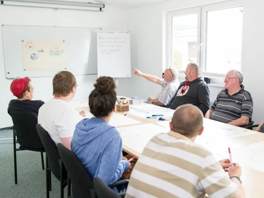 Im Besprechungsraum sitzen Frauen und Männer und sehen auf ein Whiteboard - ein Mann streckt seine Hand aus um zu erklären