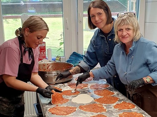 Drei Frauen bereiten gemeinsam Pizzen vor. Sie stehen um einen großen Metalltisch und bestreichen mehrere Pizzateige mit Tomatensauce, während sie Schutzhandschuhe tragen.