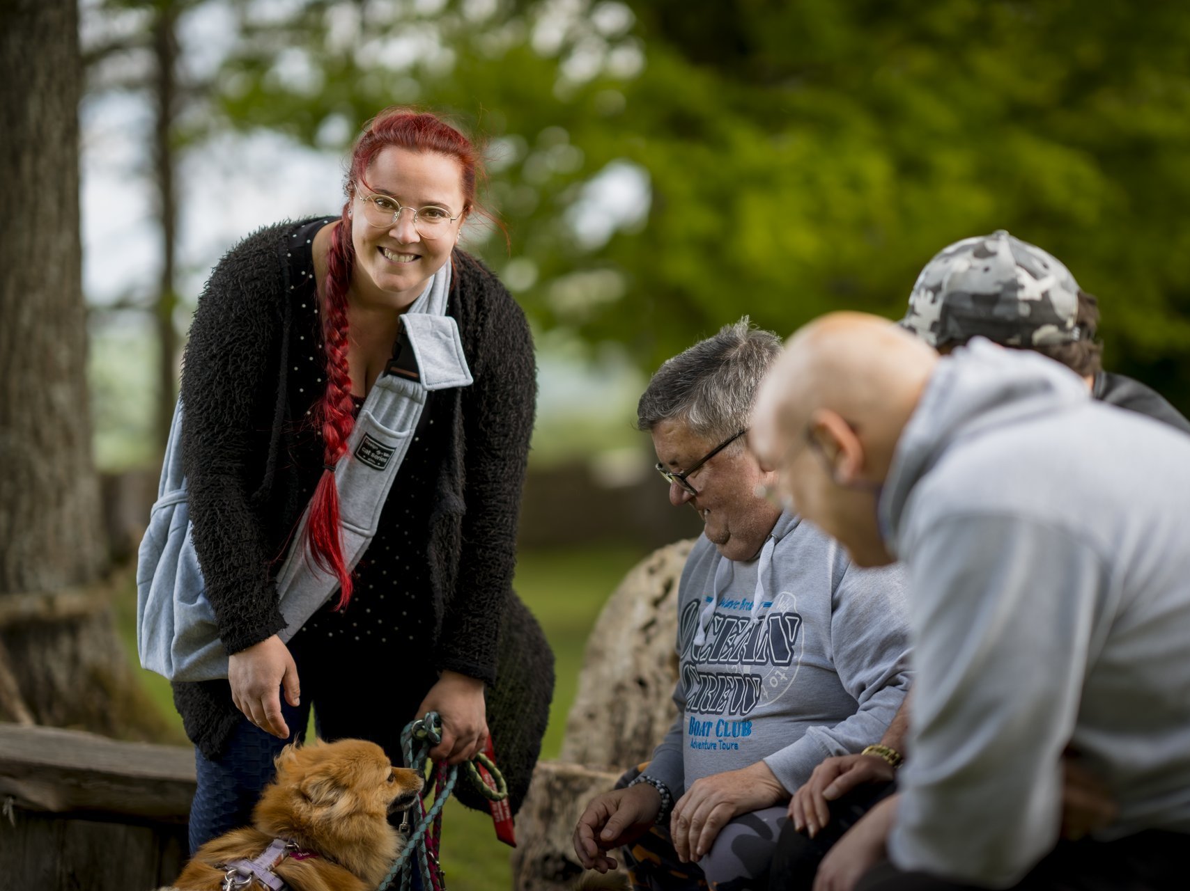 Rothaarige Frau lächelt in die Kamera, rechts sitzen drei Männer auf einer Holzbank und spielen mit einem kleinen Hund.
