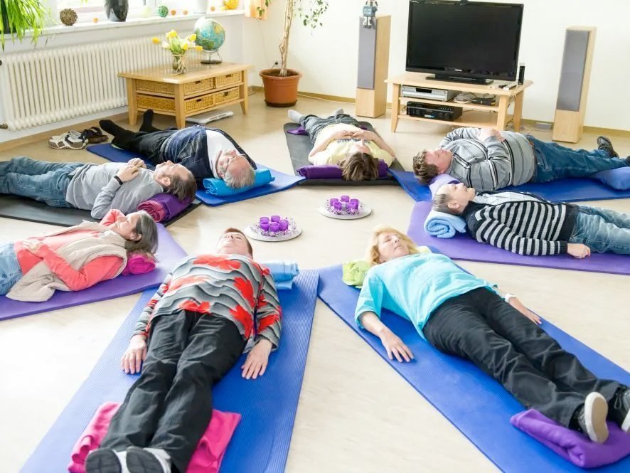 Eine Gruppe Frauen und Männer liegen auf Yoga-Matten im Kreis, haben die Augen geschlossen und sehen entspannt aus
