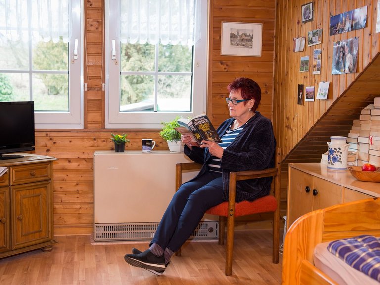Eine ältere Frau sitzt vor der Heizung ihres gemütlichen Zimmers und liest das Buch: "Die linke Hand des Bösen - von Wolfgang Burger"