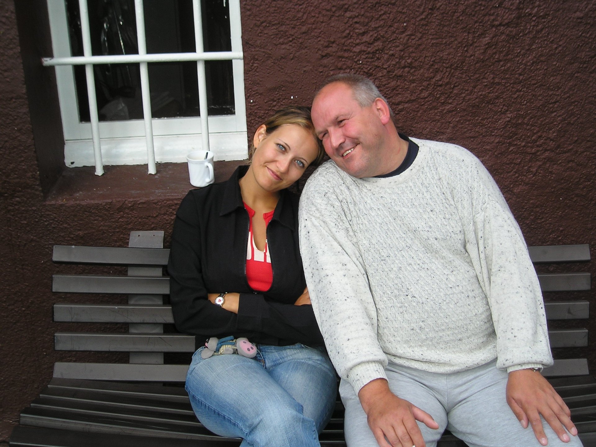 Junge Frau mit strahlend blauen Augen und ein Mann mit kurzen grauen Haaren sitzen Kopf an Kopf gelehnt auf einer Bank